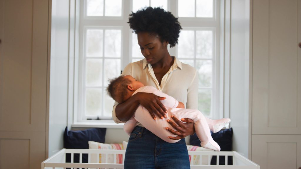 Mother holding a newborn in a bright nursery beside a white crib, looking down at the baby.