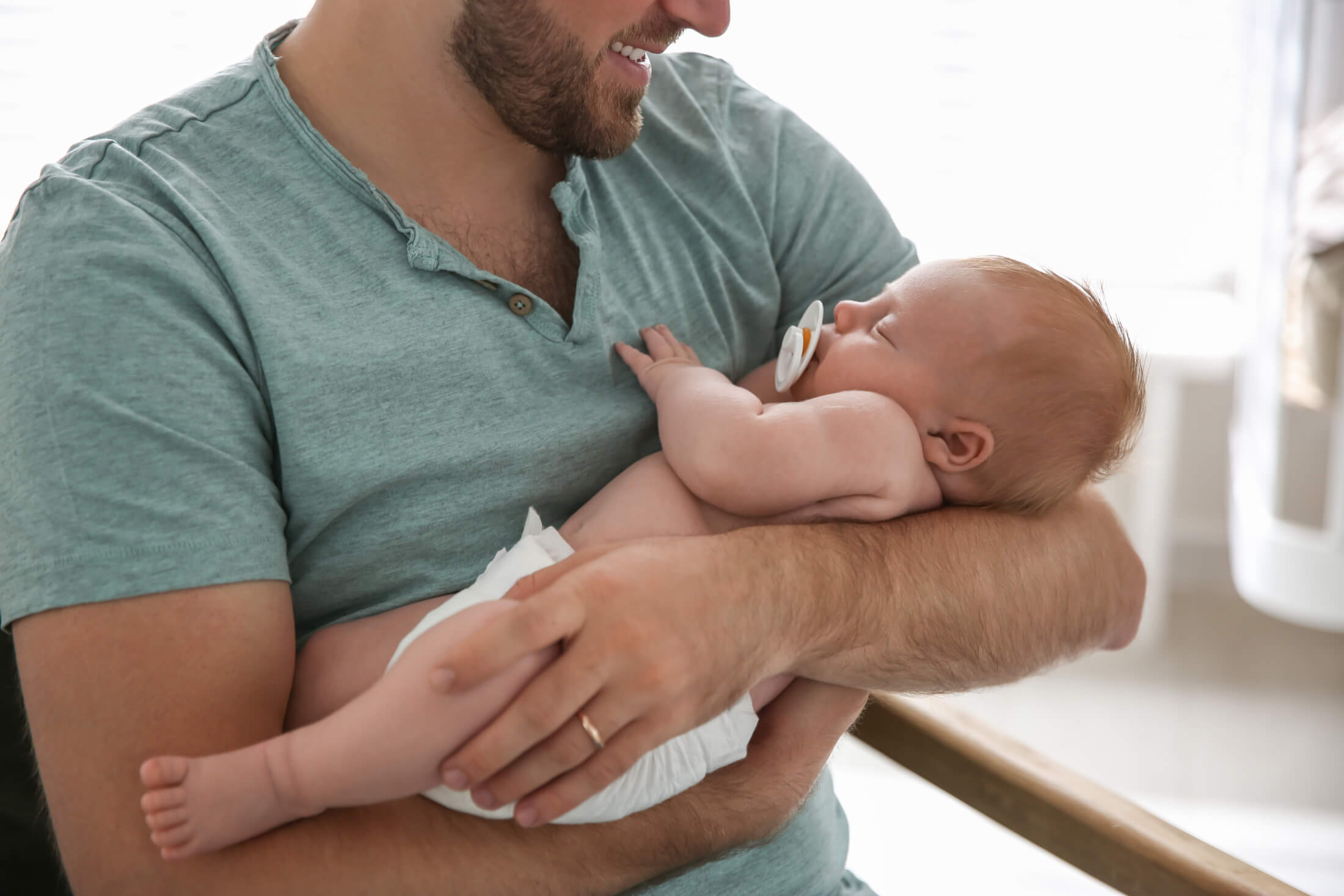Bearded man in a teal shirt holding a sleeping newborn baby with a pacifier.