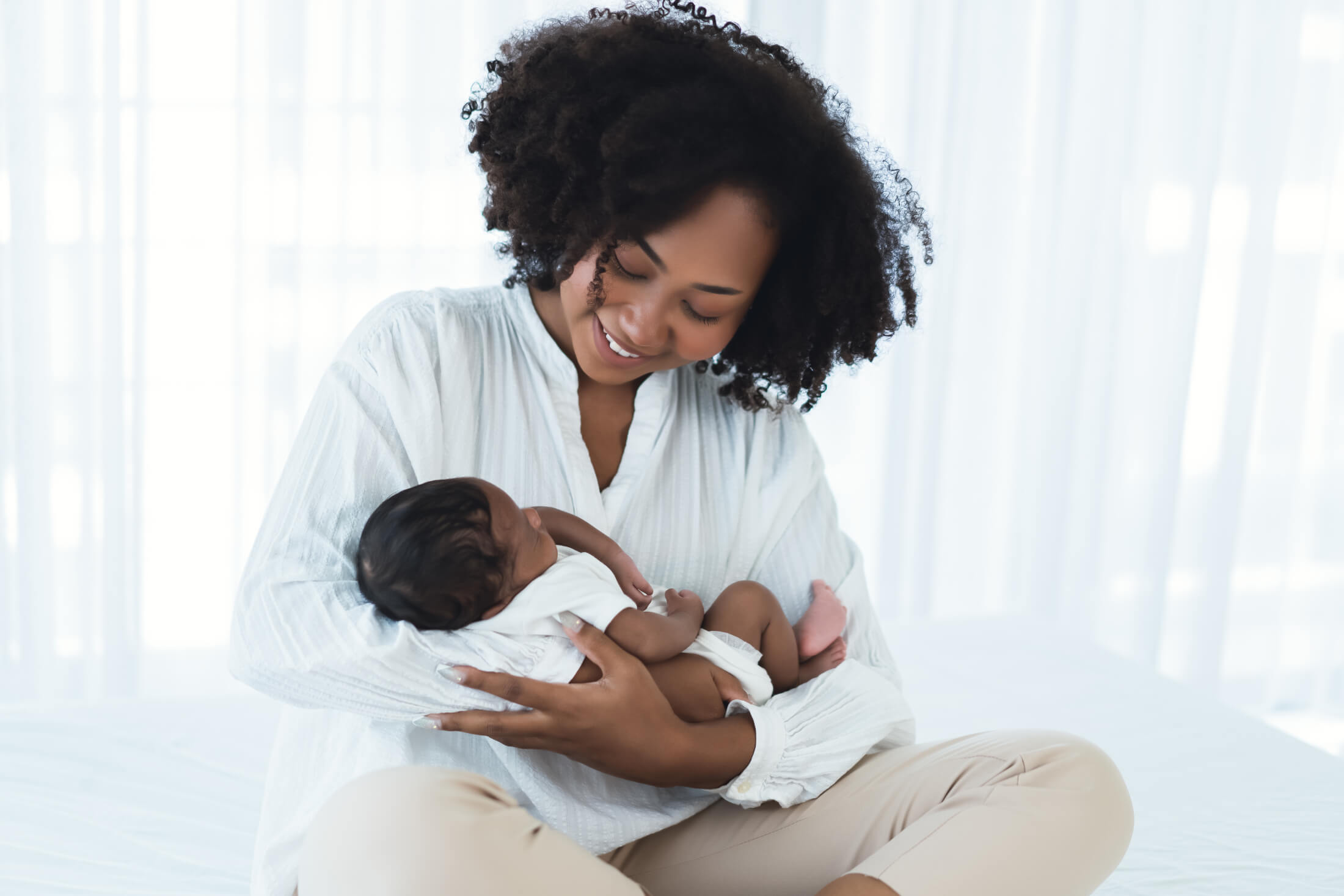 Mother gently holding a newborn baby in her arms, smiling in a bright, airy room with white curtains