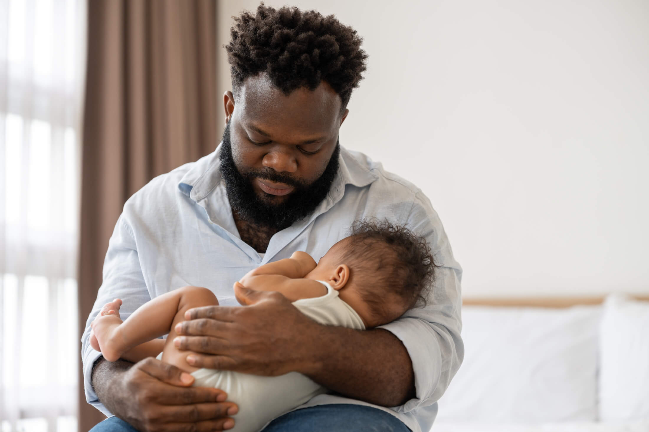 Bearded father in a light blue shirt cradling a newborn baby close to his chest, looking down lovingly.